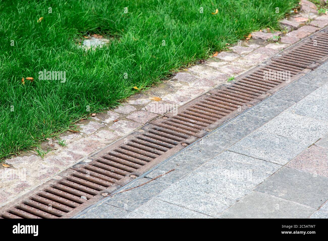 rust drainage grate of a storm system on a pedestrian sidewalk made of stone tiles in a park near a green grass. Stock Photo