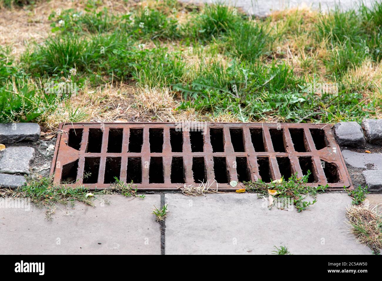 storm hatch sewage with a rusty grill on the edge of a pedestrian stone walkway overgrown with weeds in the park. Stock Photo