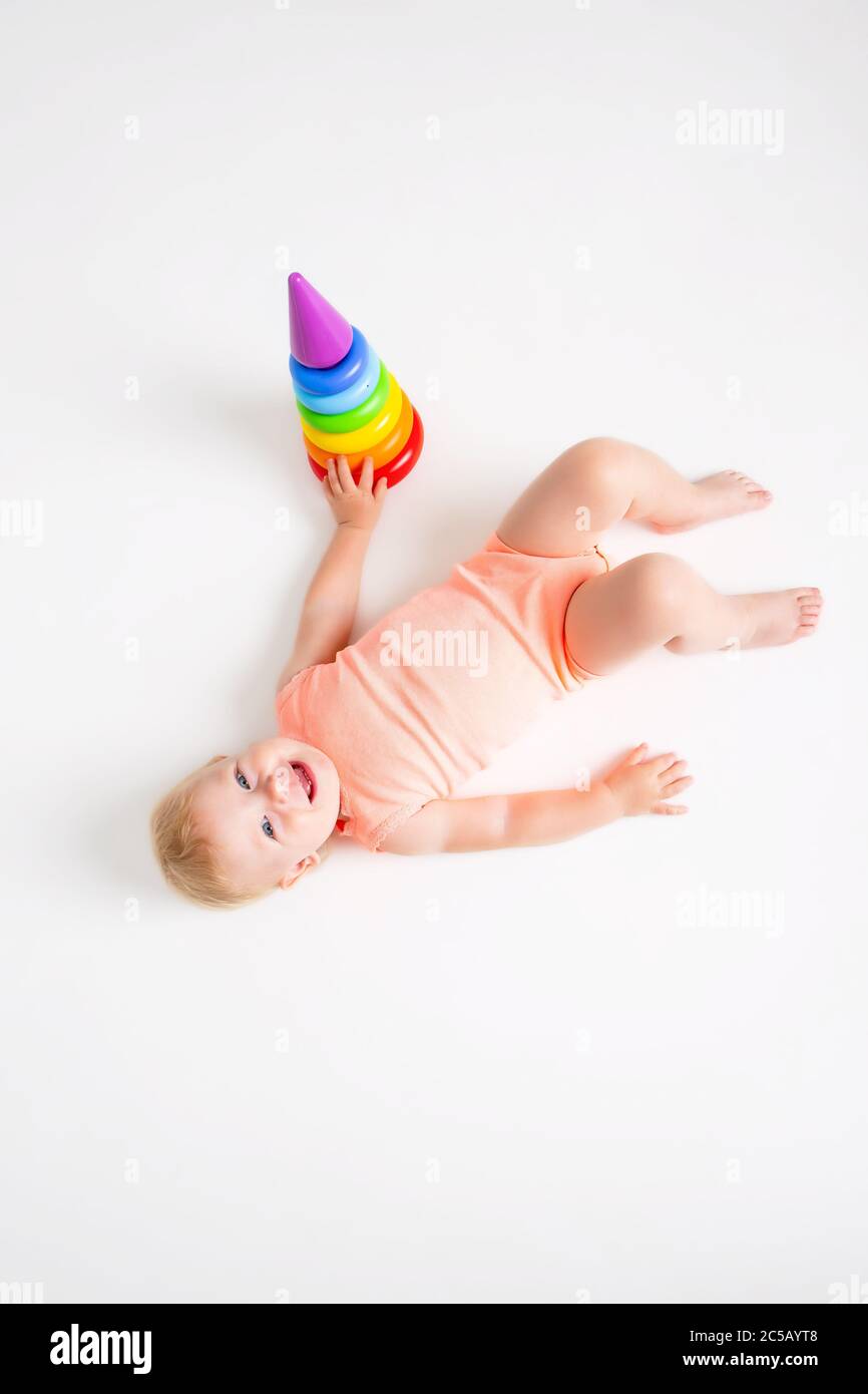 Toddler girl in orange bodysuit lies next to a multi-colored pyramid on a white background, view from above, space for text Stock Photo