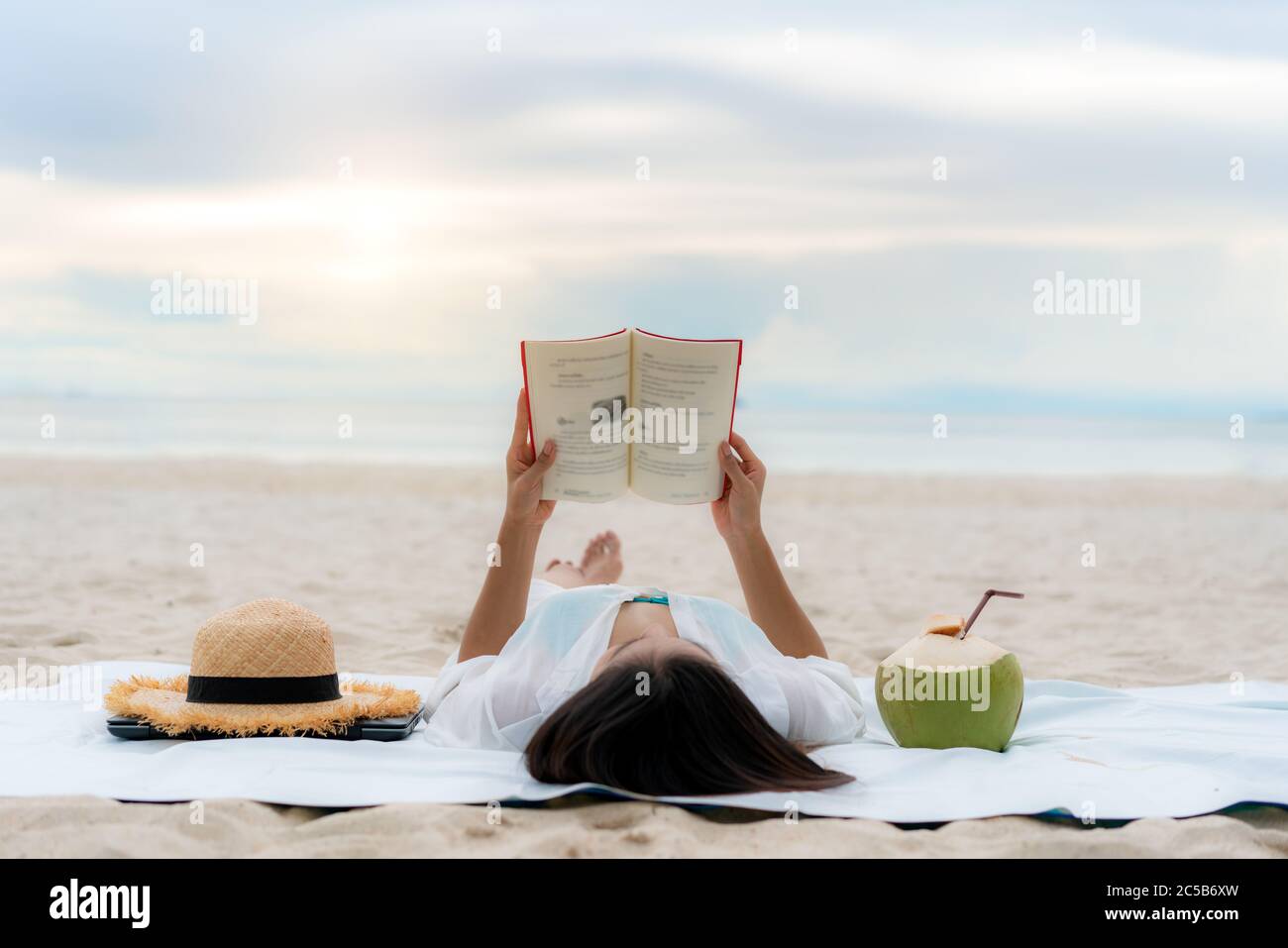 Young Asian woman lying on a tropical summer beach sea for relax reading book with coconut juice beside her. Summer, holidays, vacation and happy peop Stock Photo