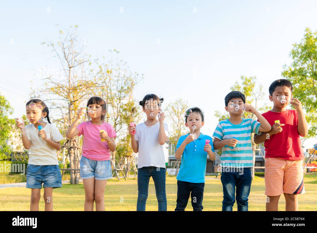 Large group of happy Asian smiling kindergarten kids friends playing blowing bubbles together in the park on the green grass on sunny summer day. Stock Photo