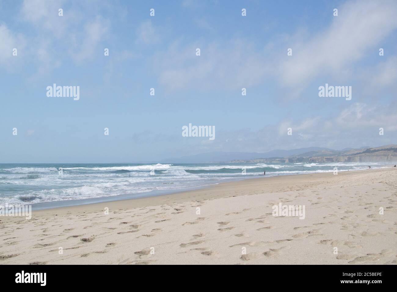 San Gregorio State Beach, blue sky, white fluffy clouds, waves rolling in, pale sand with footprints, cliffs and mountains in distance. Stock Photo