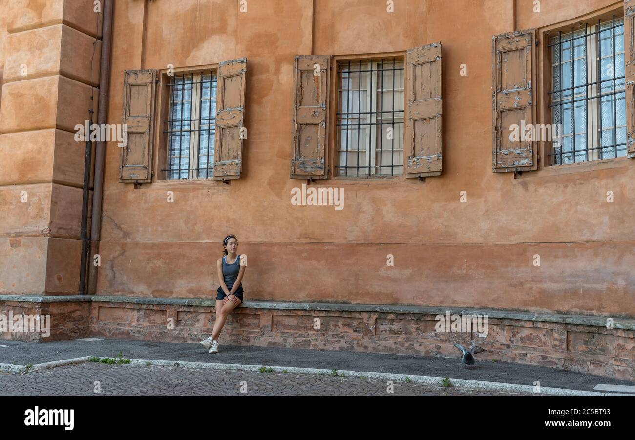 Young teen girl with headband, shorts, tanktop and sneakers sitting on brick bench under 3 windows with shutters and bars in Bologna Italy Stock Photo