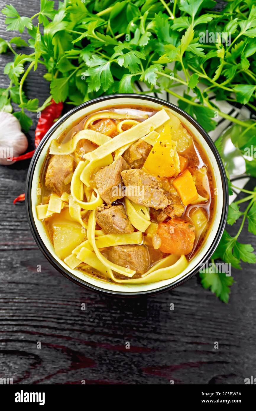 Central Asian national dish Lagman of meat, noodles and vegetables in bowl on a napkin, garlic, parsley on wooden board background from above Stock Photo