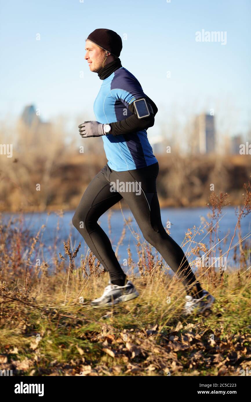 Running man jogging in autumn listening to music on smart phone. Runner  training in warm outfit on cold day. Fit male fitness athlete model  training outdoor in fall. Full body length of