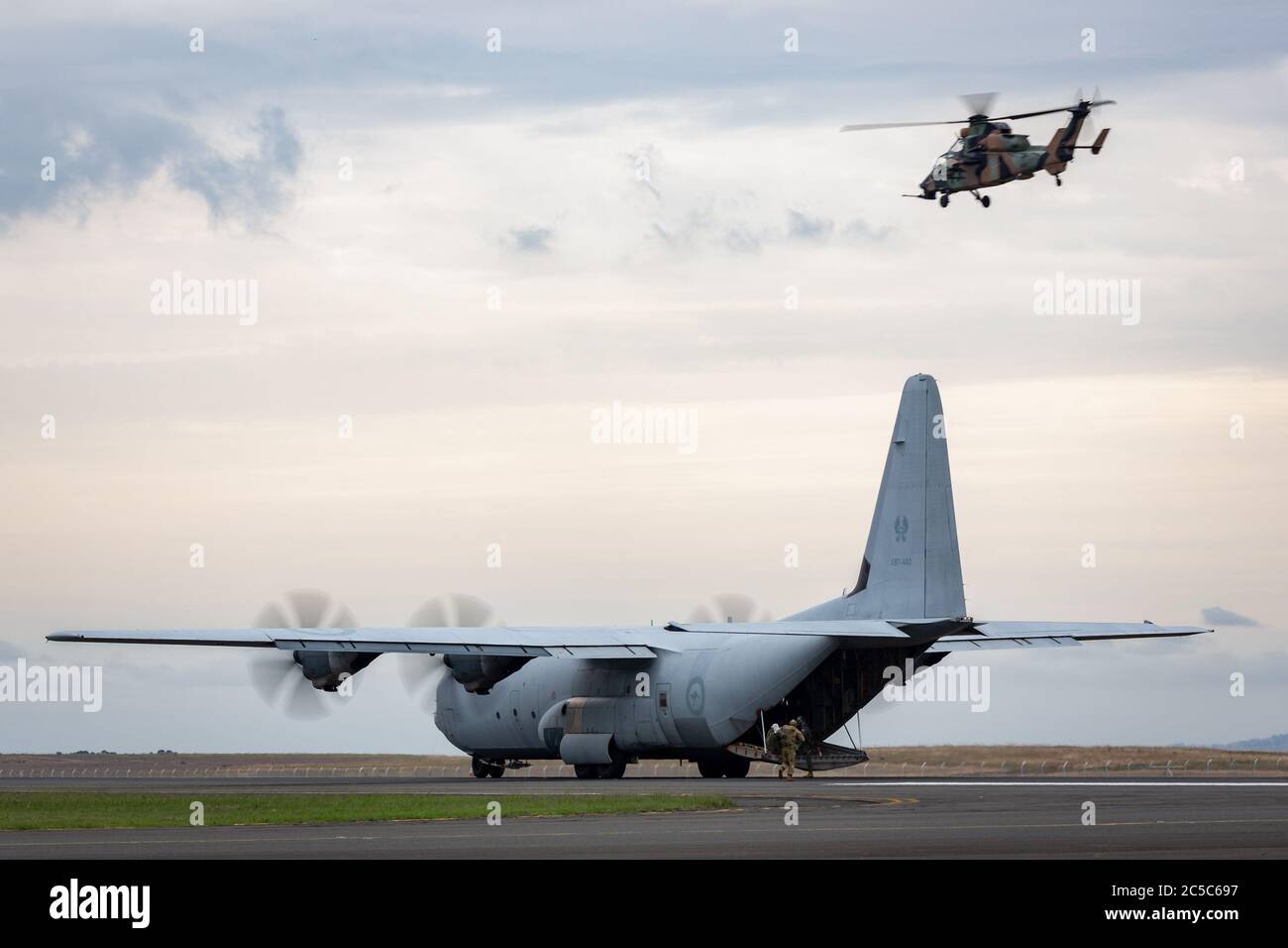 Royal Australian Air Force Lockheed Martin C-130J Hercules military cargo aircraft on the runway with an Army Eurocpter Tiger ARH helicopter flying ab Stock Photo