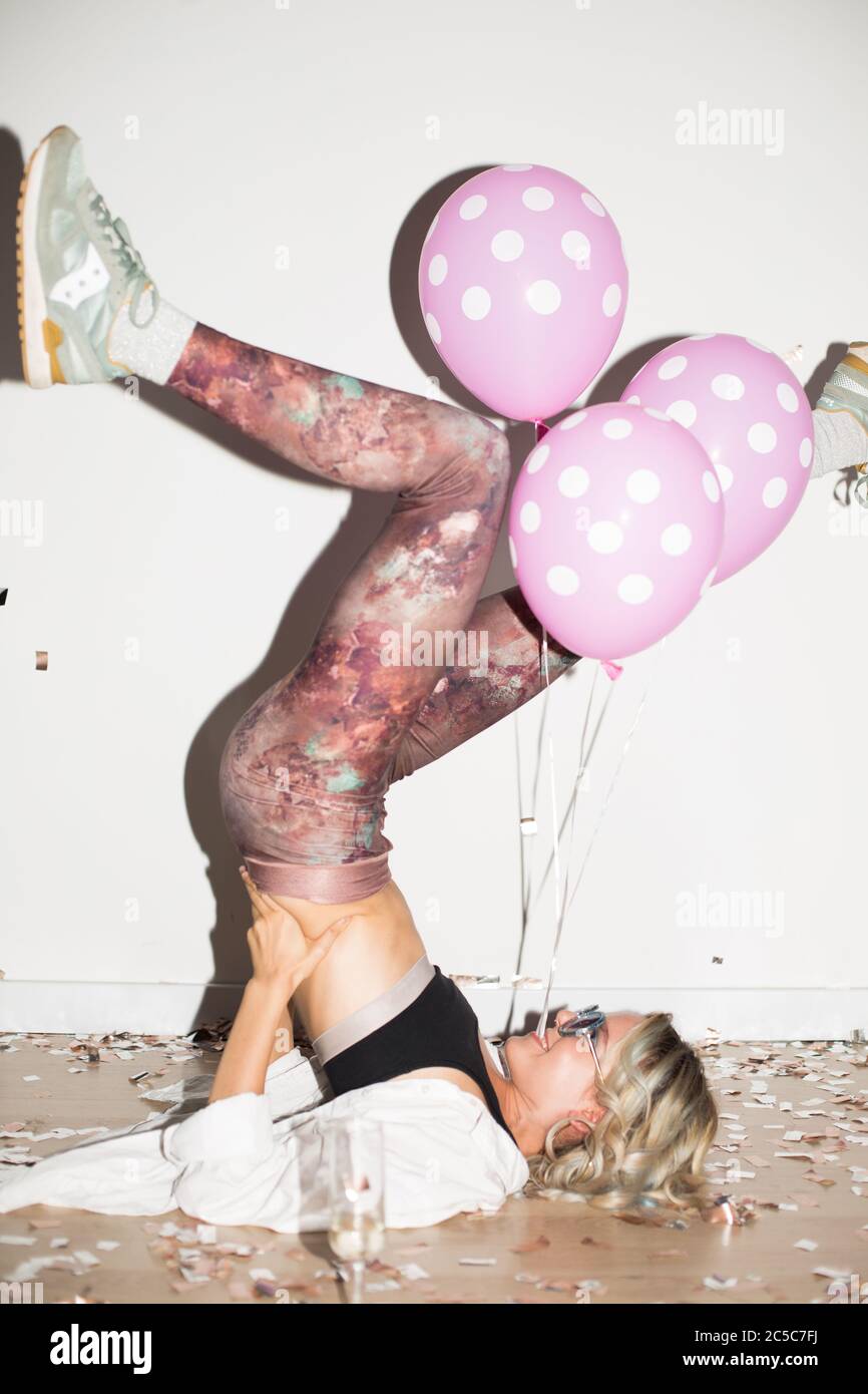 Joyful girl in sunglasses with pink balloons near lying on floor while lifting legs up with confetti around over white background Stock Photo