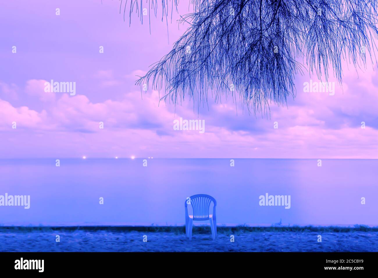 Calm seascape and empty chair on a beach at dusk, spotlights of fishing boats off the coast, dramatic purple clouds over an ocean. Long exposure. Stock Photo