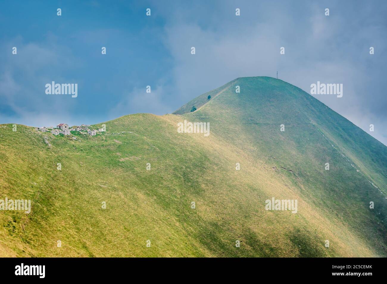 Green hill with steep slopes, an old abandoned stone farm on the ridge. Hiking trail leading to a peak with a cross, Mount Crocione, Italy. Stock Photo