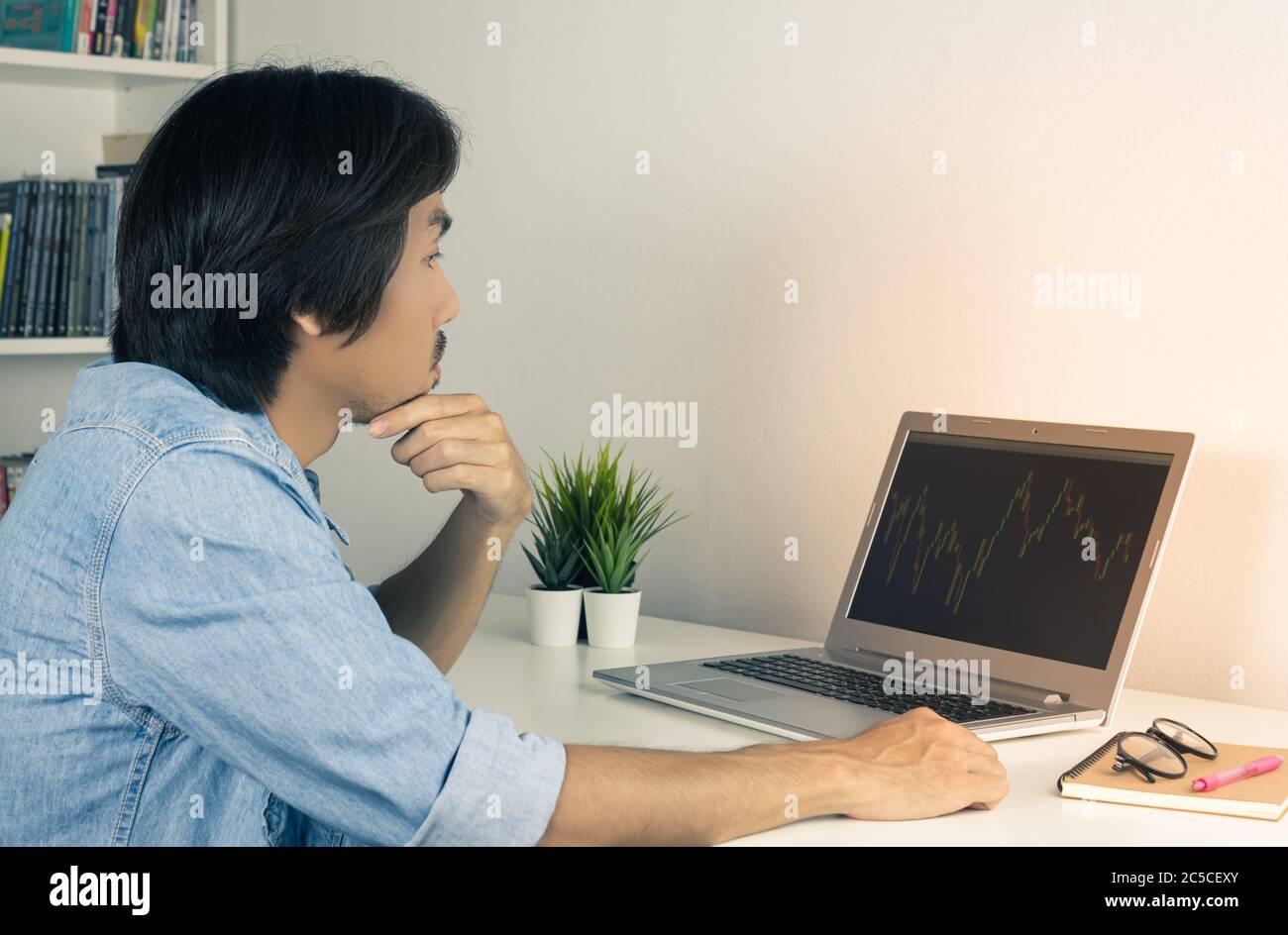 Asian Forex Trader or Investor or Businessman Wear Jean Shirt Trading and Looking Forex Charts or Stock Charts in Laptop Monitor. Serious Casual Busin Stock Photo