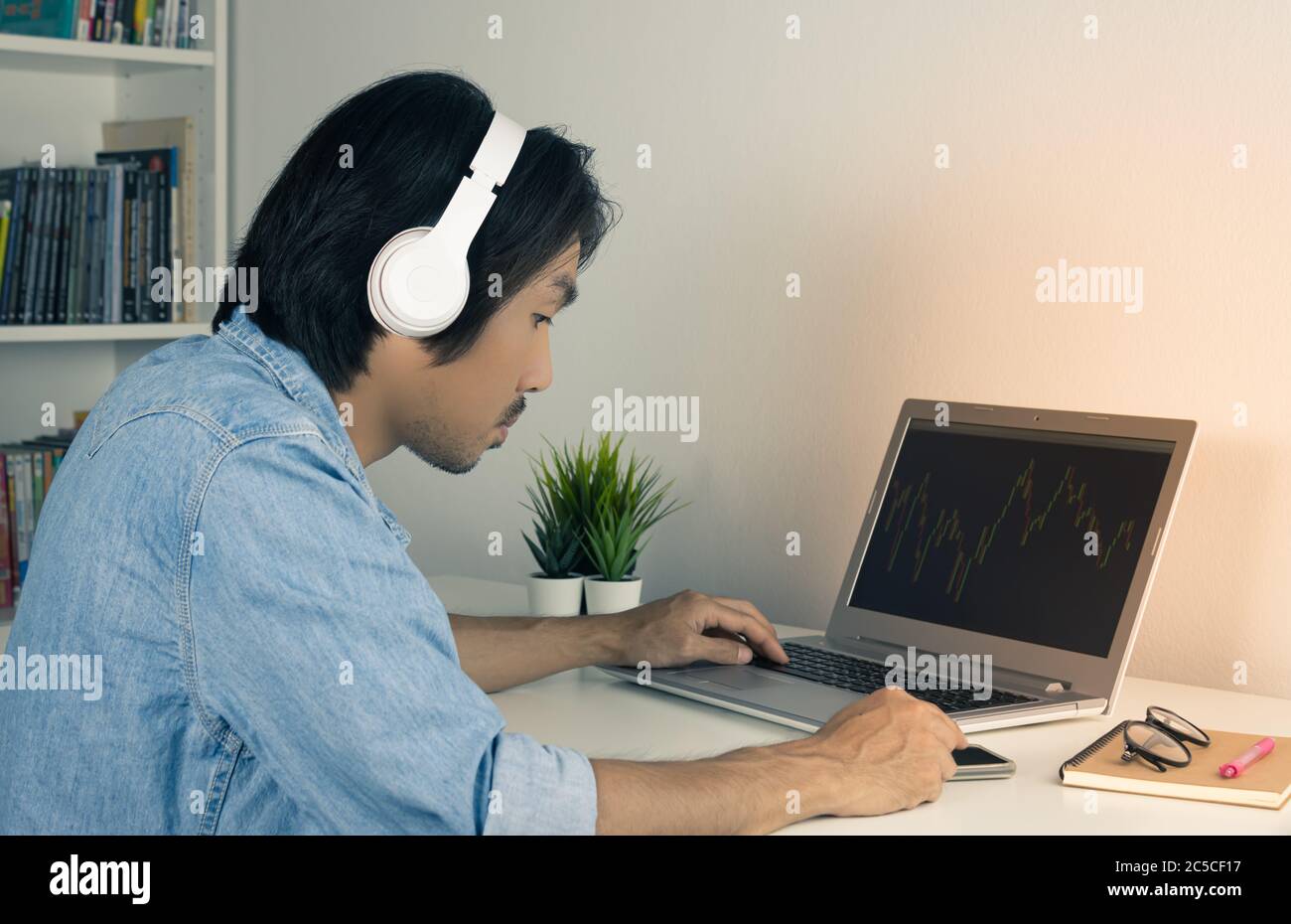 Asian Forex Trader or Investor or Businessman Wear Jean Shirt and Headphone Trading Forex Charts or Stock Charts in Laptop and Touch Smartphone. Casua Stock Photo