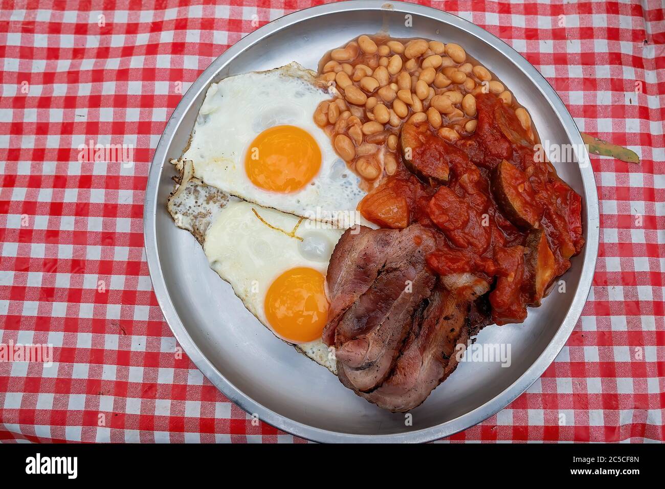 Bacon eggs tomato and beans served on a tin plate on a checked tablecloth at a campfire bush breakfast on an Australian outback tourist resort Stock Photo
