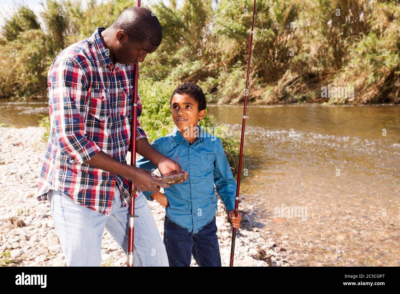 African man and little boy standing near river and holding fish on fishing rod Stock Photo