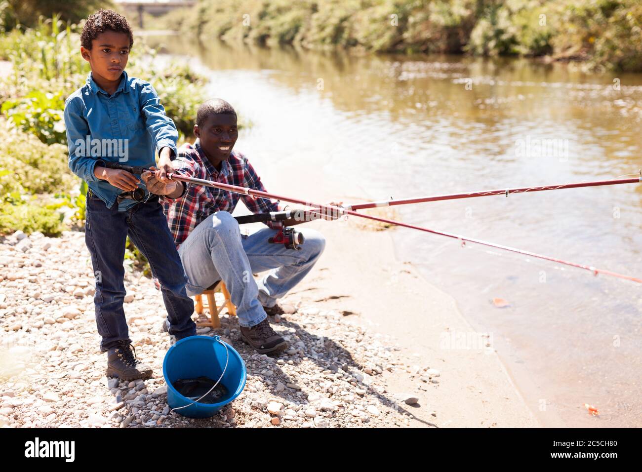 Positive African man and little boy standing near river and fishing with rods Stock Photo