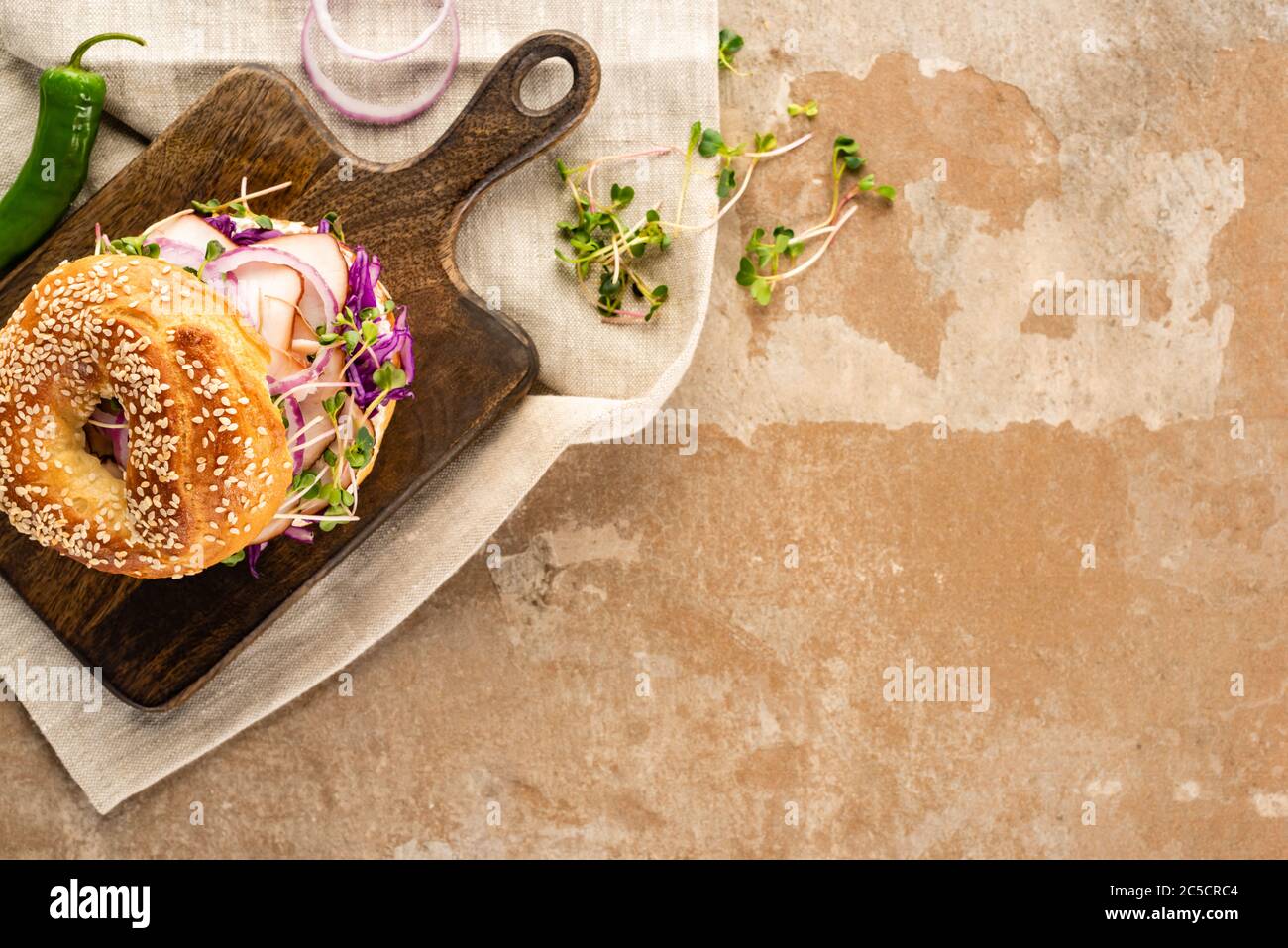 top view of fresh delicious bagel with meat, red onion and sprouts on wooden cutting board on napkin with jalapenos on aged beige surface Stock Photo