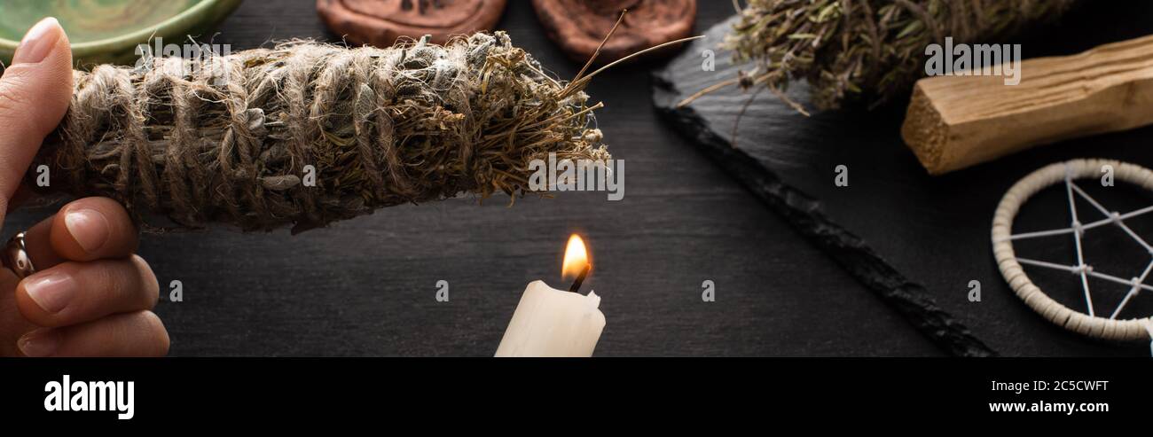 Cropped view of shaman holding smudge stick and candle near witchcraft on black wooden surface, panoramic shot Stock Photo
