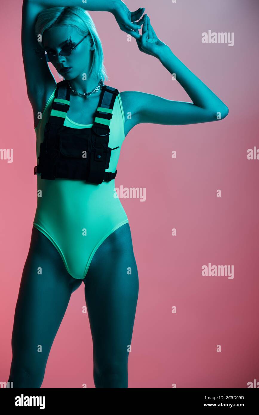 fashionable girl in futuristic leotard and fire-shaped sunglasses posing on pink in blue light Stock Photo