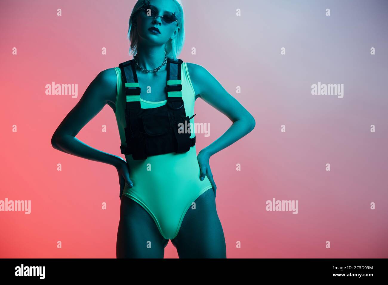 fashionable model in futuristic bodysuit and fire-shaped sunglasses posing on pink in blue light Stock Photo