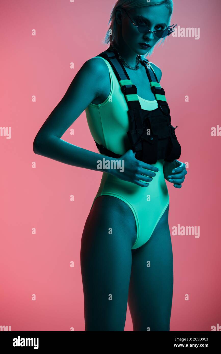 attractive fashionable girl in futuristic bodysuit and fire-shaped sunglasses posing on pink in blue light Stock Photo