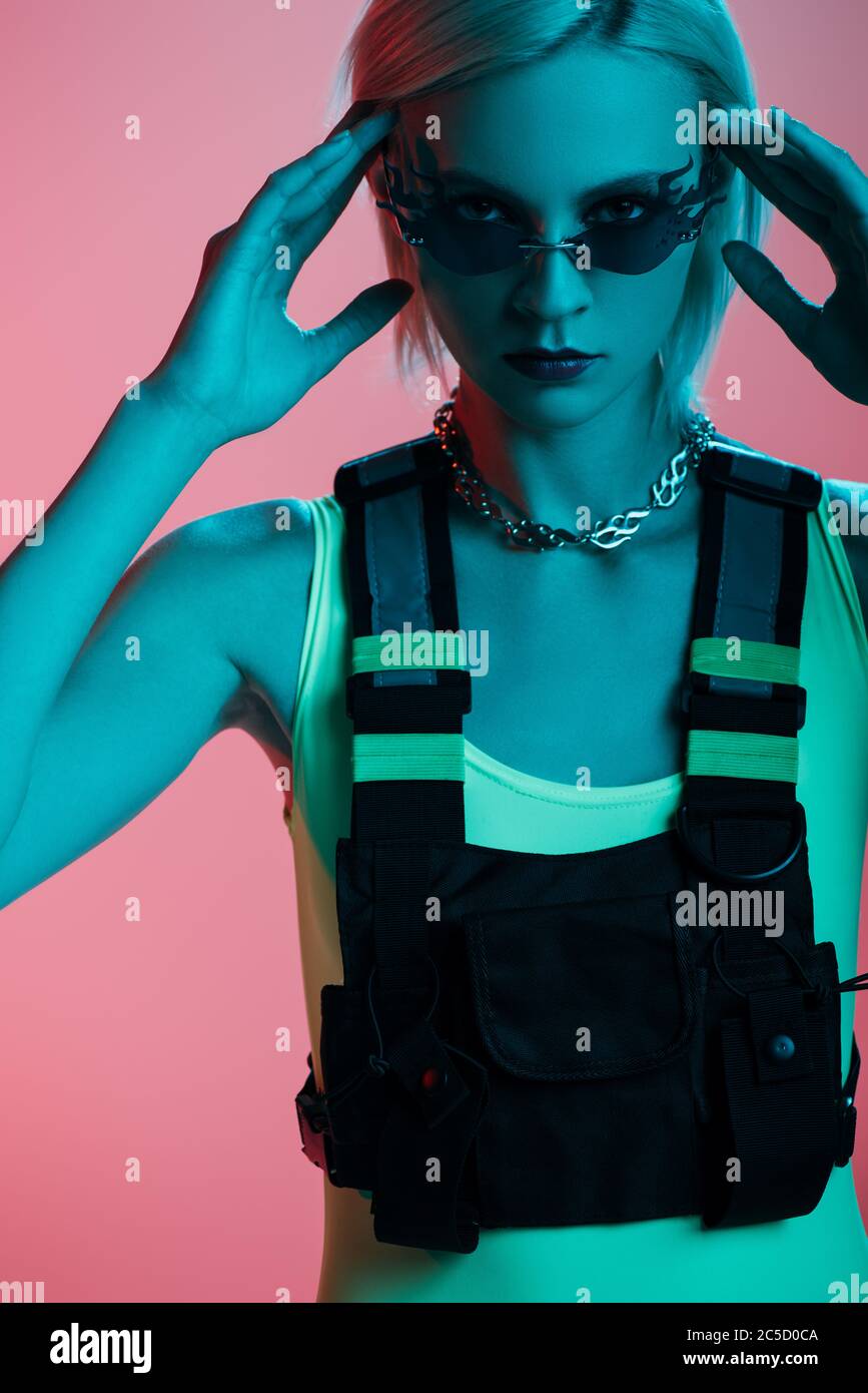 young fashionable model in futuristic bodysuit and fire-shaped sunglasses posing on pink in blue light Stock Photo