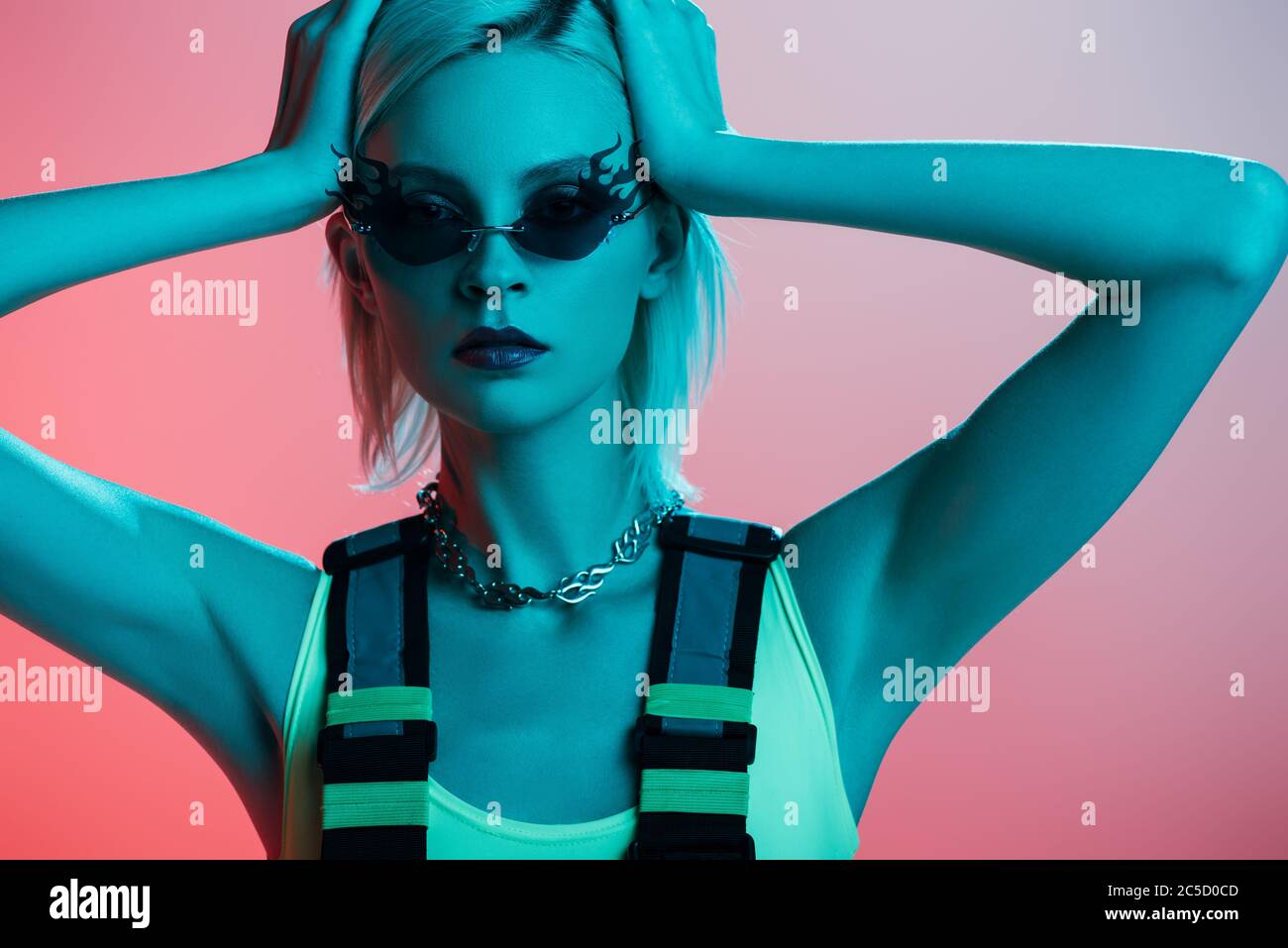 fashionable blonde model in futuristic bodysuit and fire-shaped sunglasses posing on pink in blue light Stock Photo
