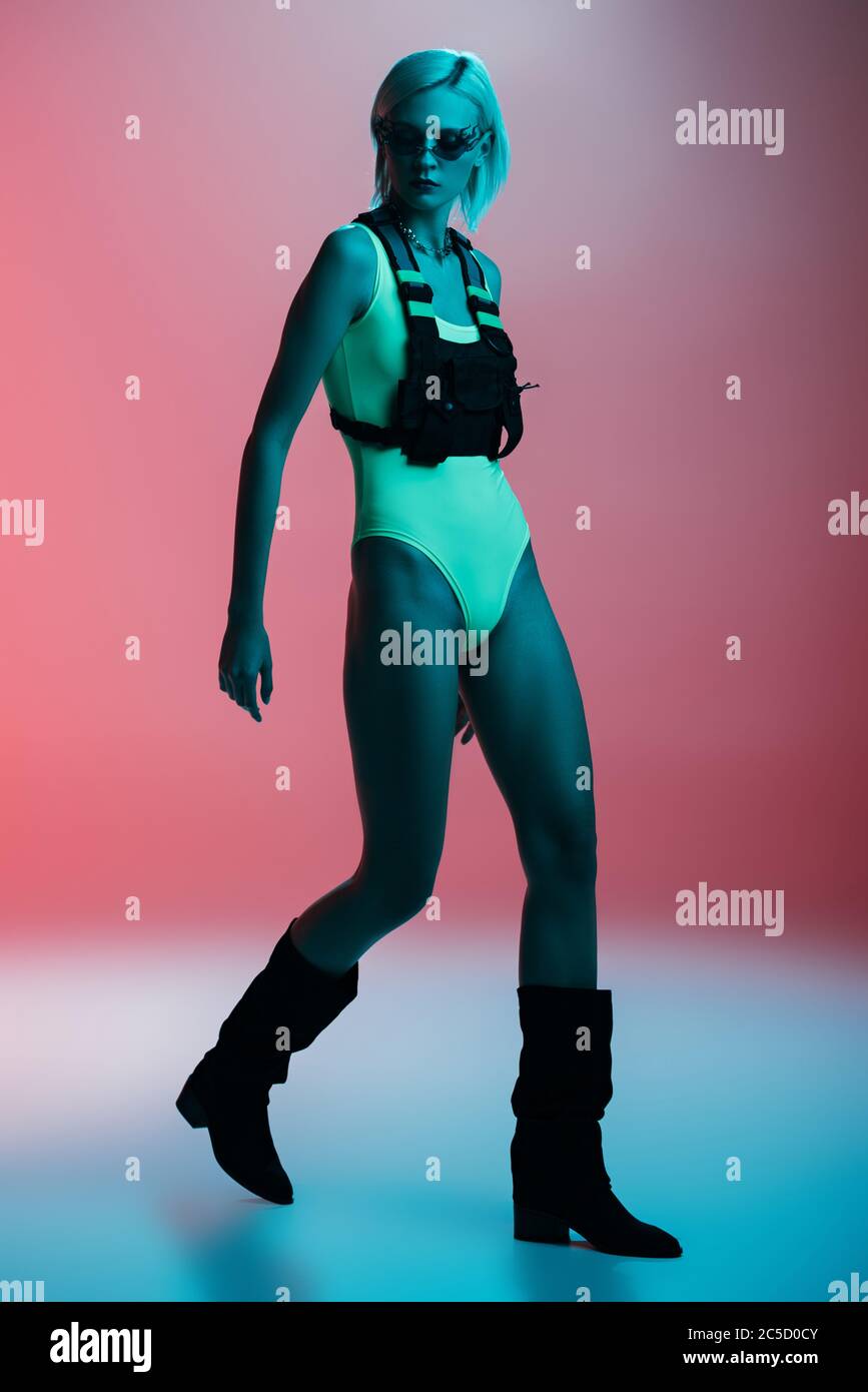 attractive fashionable model in futuristic bodysuit and fire-shaped sunglasses posing on pink in blue light Stock Photo