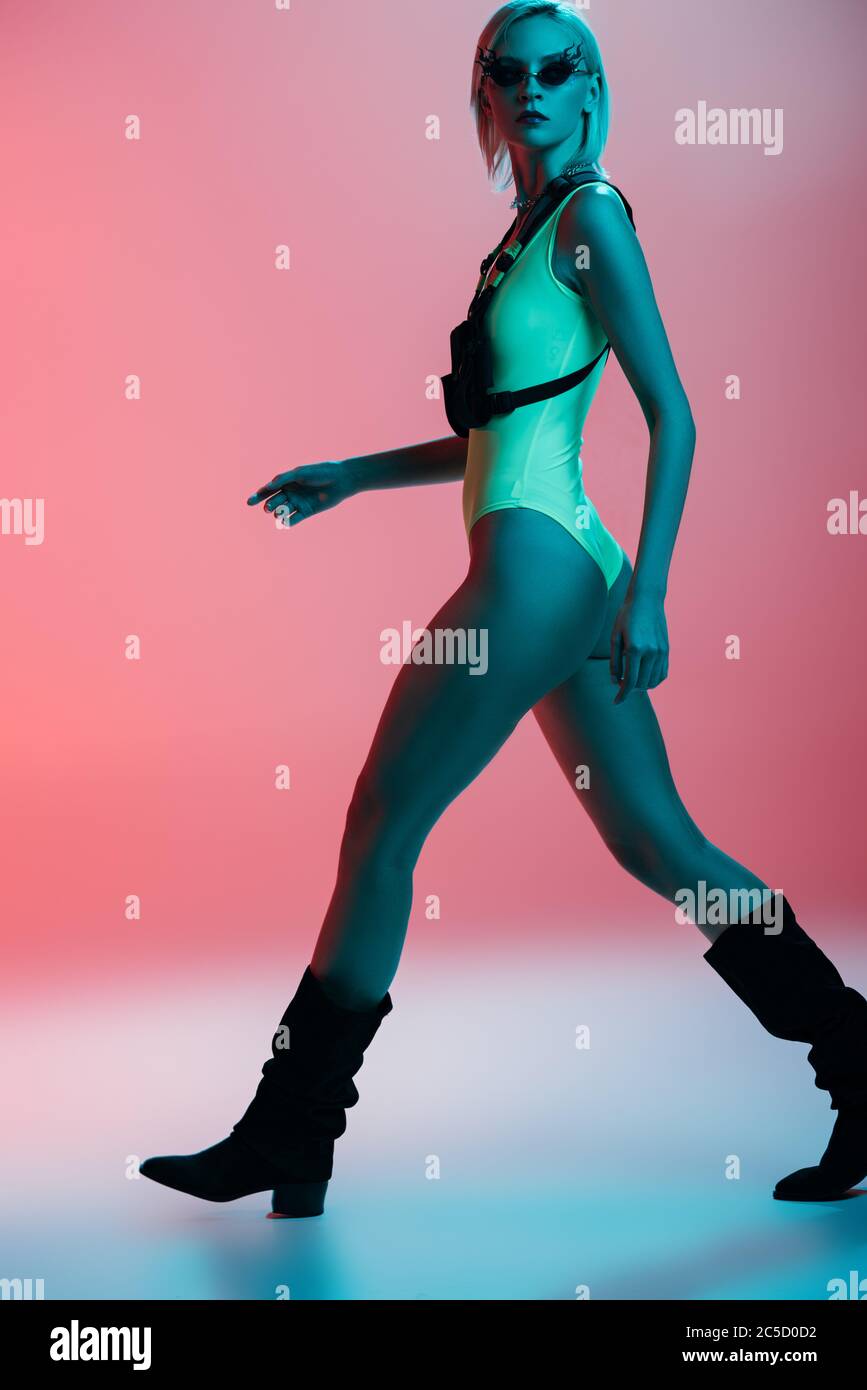 fashionable young model in futuristic leotard and fire-shaped sunglasses posing on pink in blue light Stock Photo