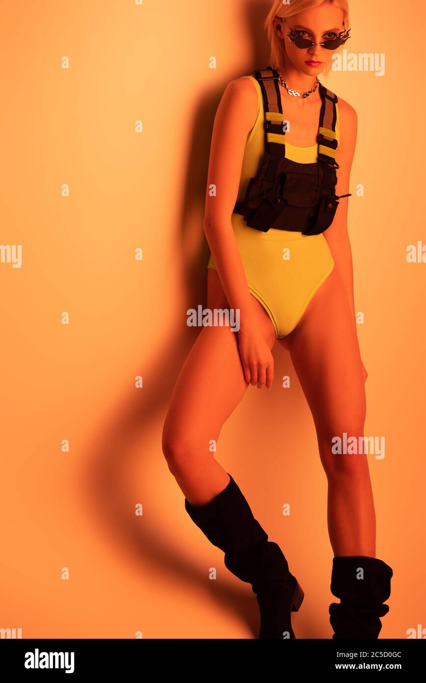 stylish futuristic girl posing in bodysuit and fire-shaped sunglasses on orange Stock Photo