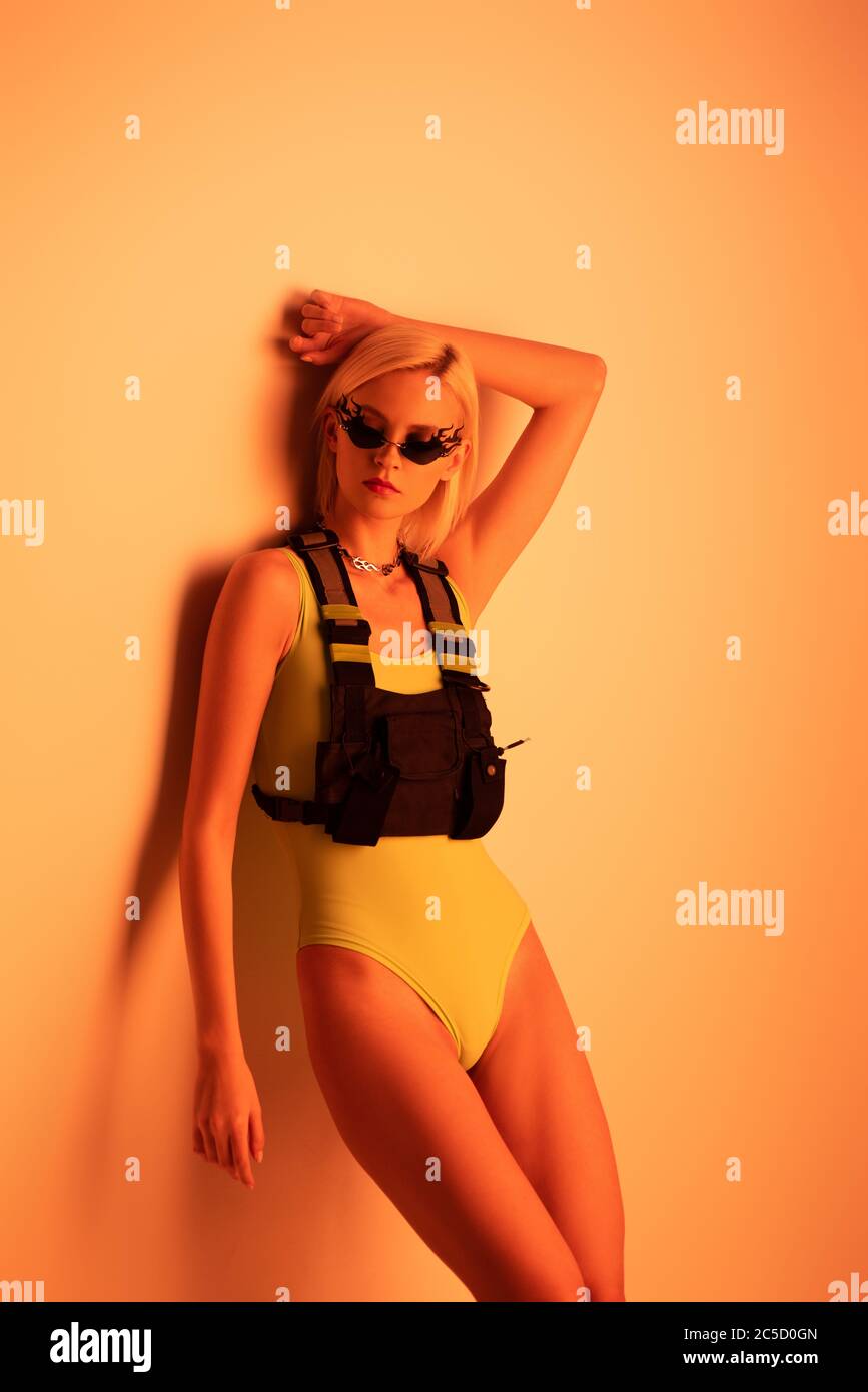 attractive futuristic girl posing in bodysuit and fire-shaped sunglasses on orange Stock Photo