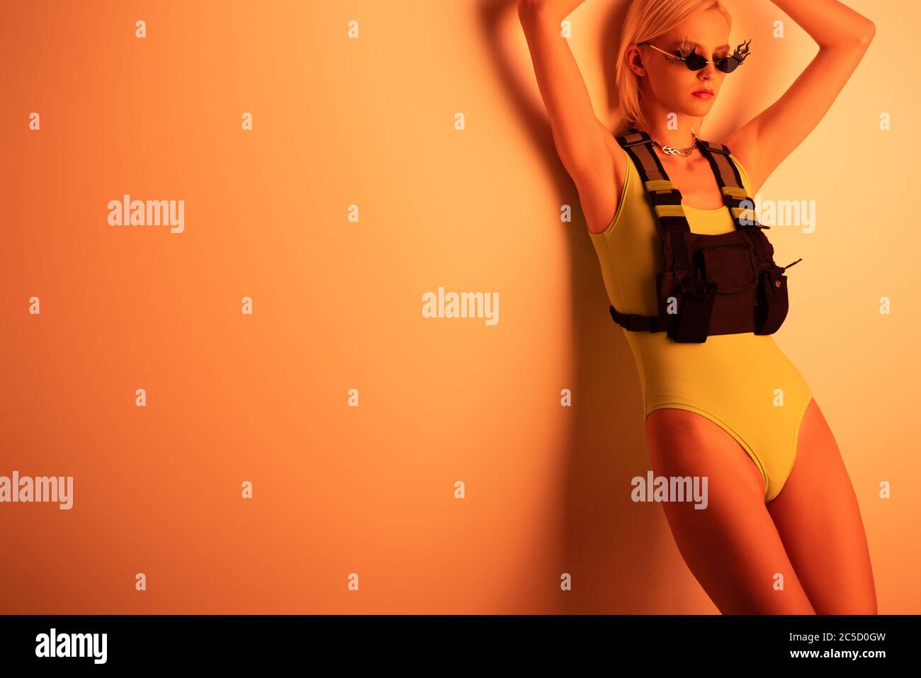 fashionable futuristic girl posing in bodysuit and fire-shaped sunglasses on orange Stock Photo