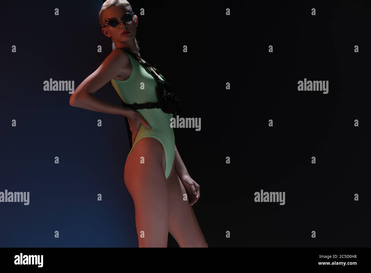 beautiful fashionable woman in futuristic bodysuit and fire-shaped sunglasses posing on dark Stock Photo