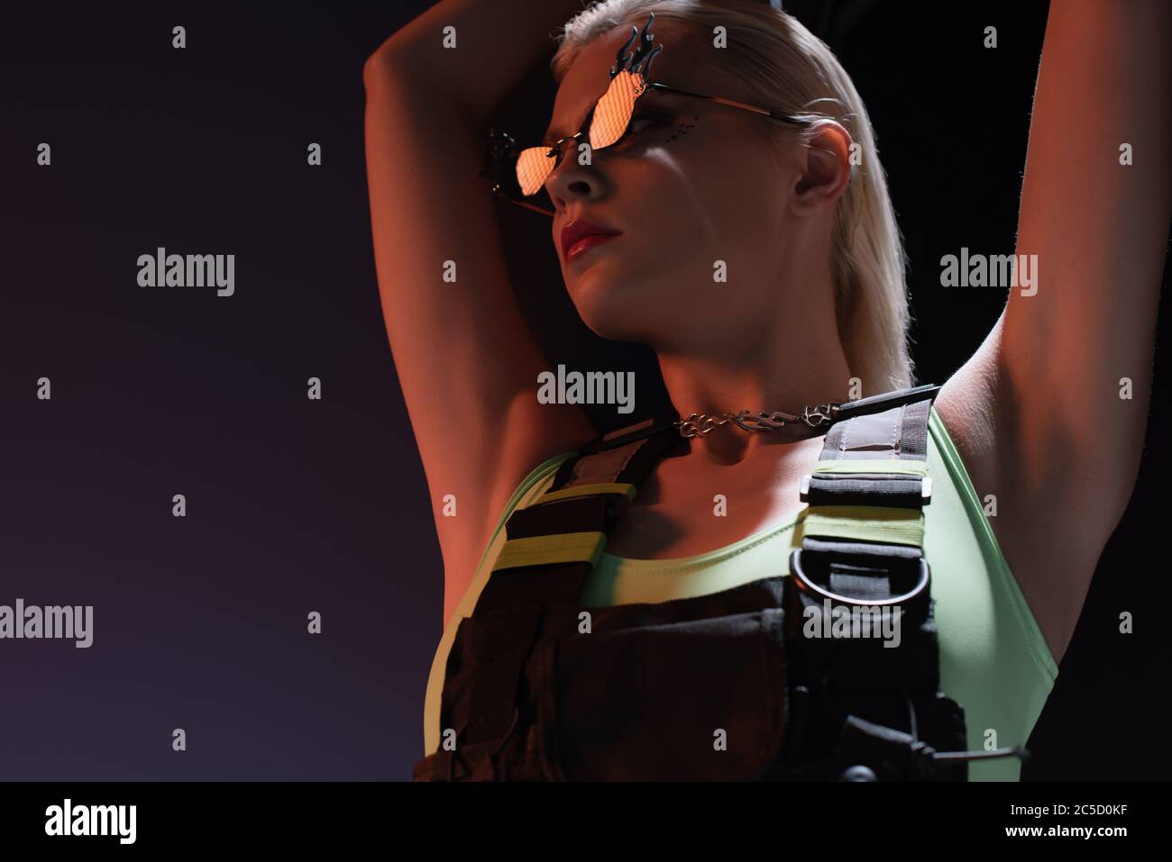 beautiful fashionable woman in futuristic bodysuit and fire-shaped sunglasses posing on dark Stock Photo