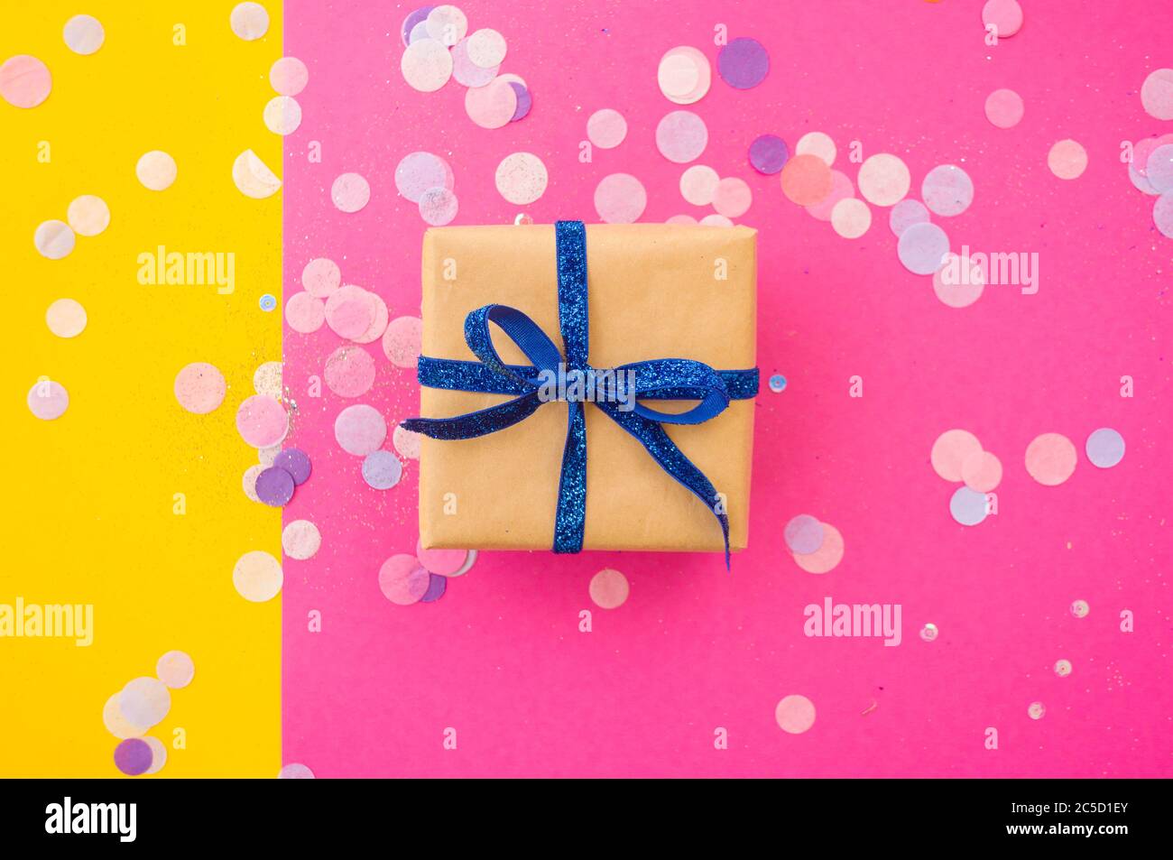 Giftbox tied with blue color ribbon on bright pink, pastel yellow background with colorful confetti and glitter. Festive background. Stock Photo