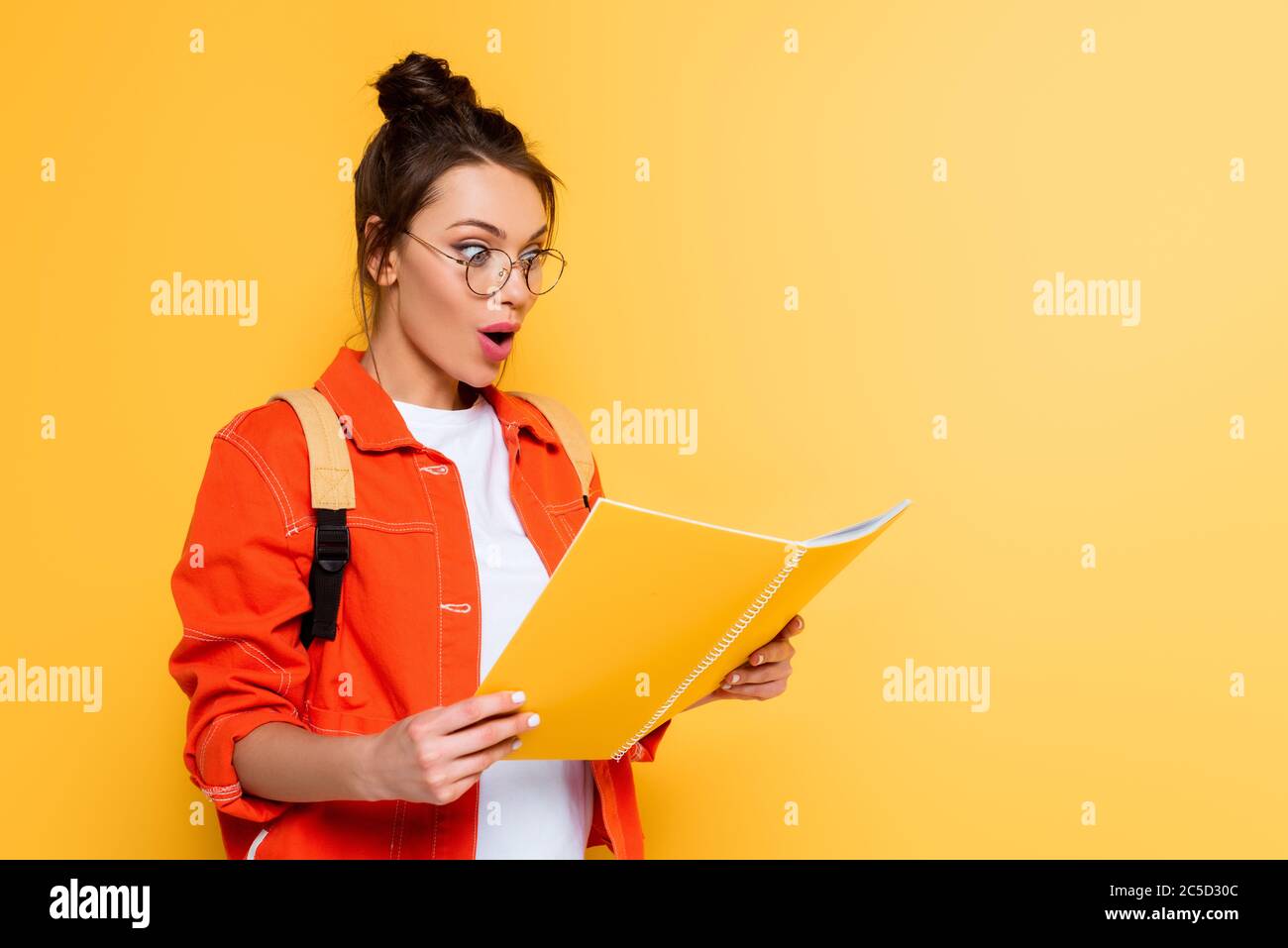 shocked student in glasses looking at copy book isolated on yellow Stock Photo