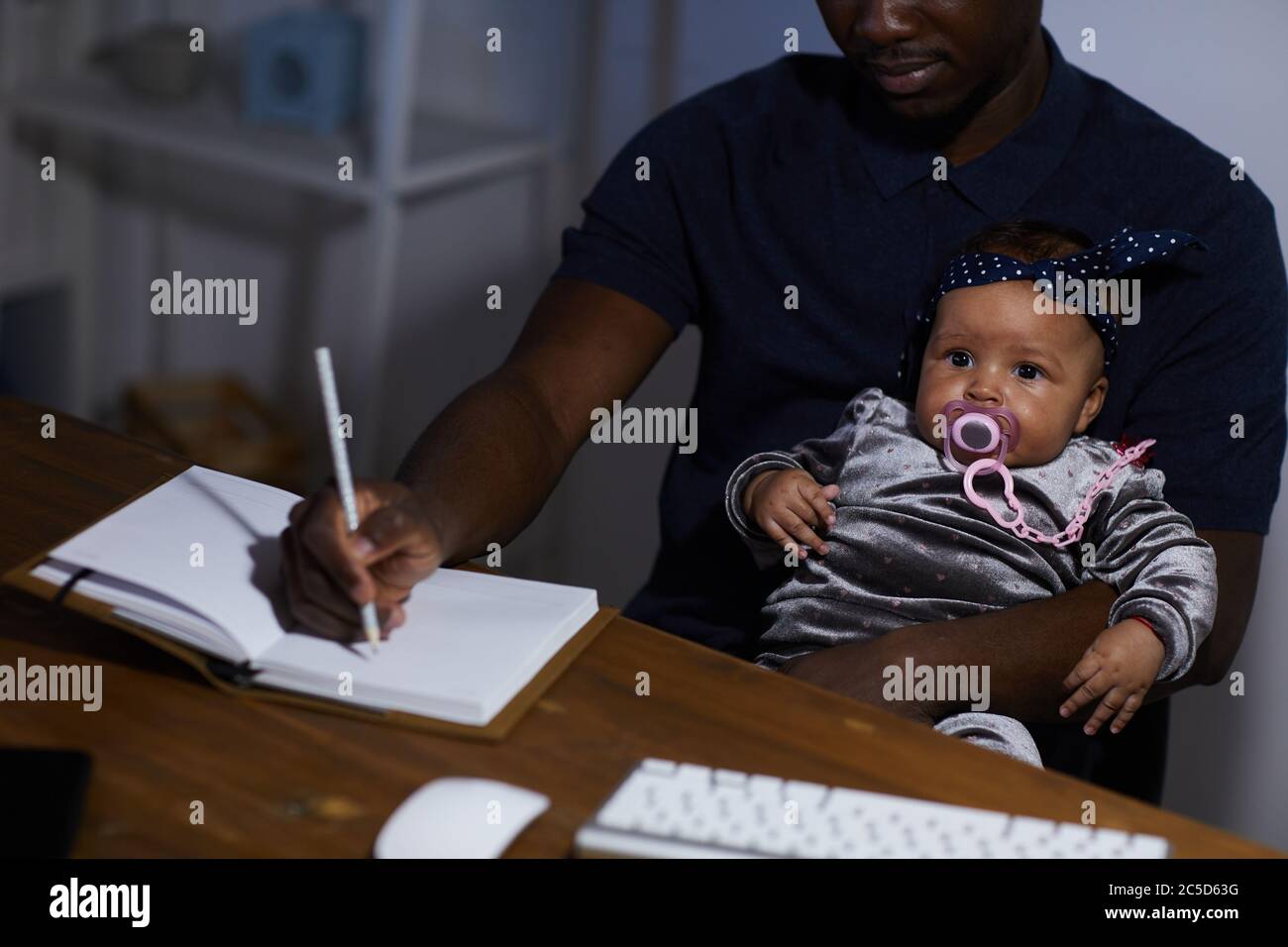 African father making notes in note pad while sitting at the table with baby on his knees Stock Photo