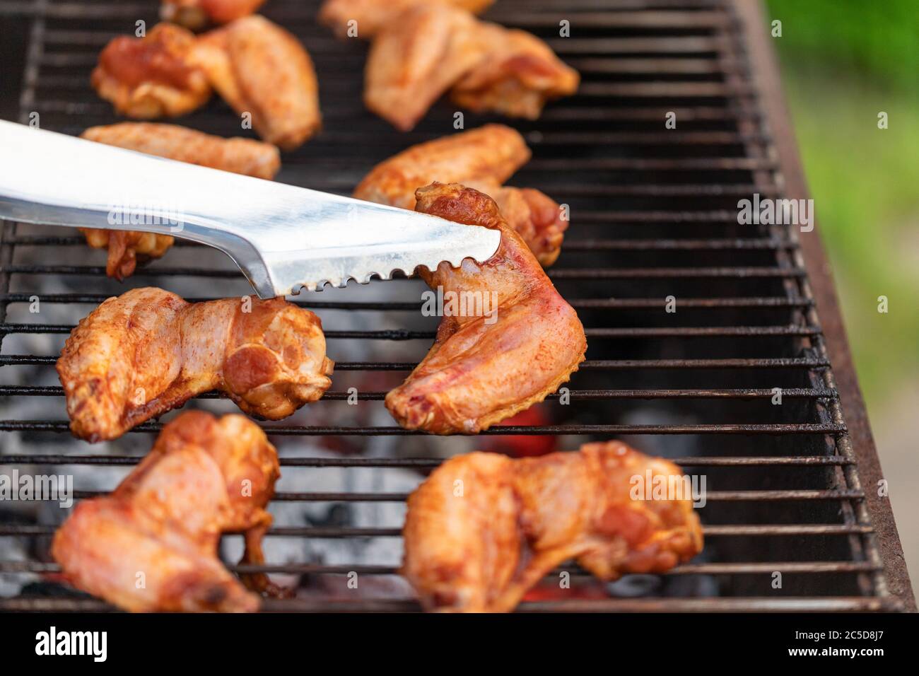 Grilled chicken wings on the grill with smoke, metal tongs to turning chicken wings. Close up, selective focus Stock Photo
