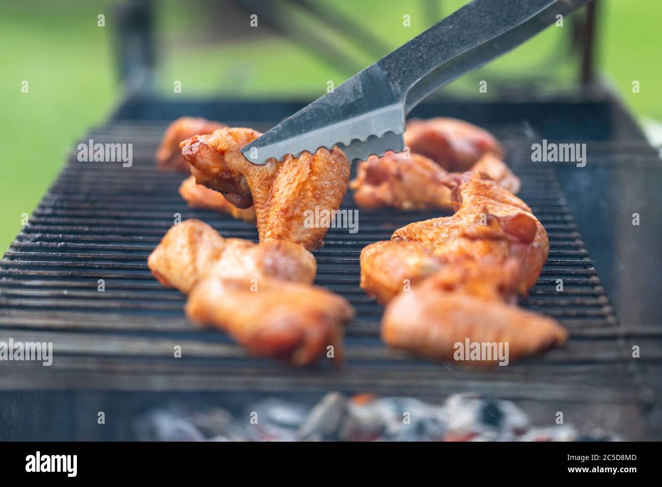 Grilled chicken wings on the grill with smoke, metal tongs to turning chicken wings. Close up, selective focus Stock Photo