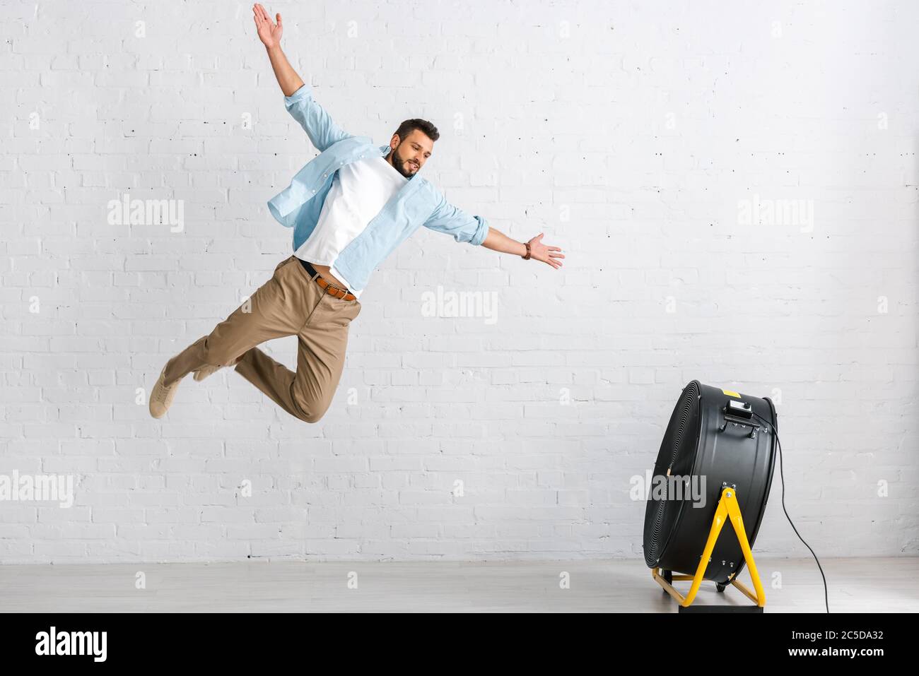 Handsome bearded man jumping near electric fan at home Stock Photo