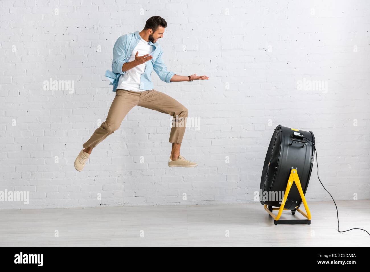 Handsome man jumping near electric fan on floor at home Stock Photo