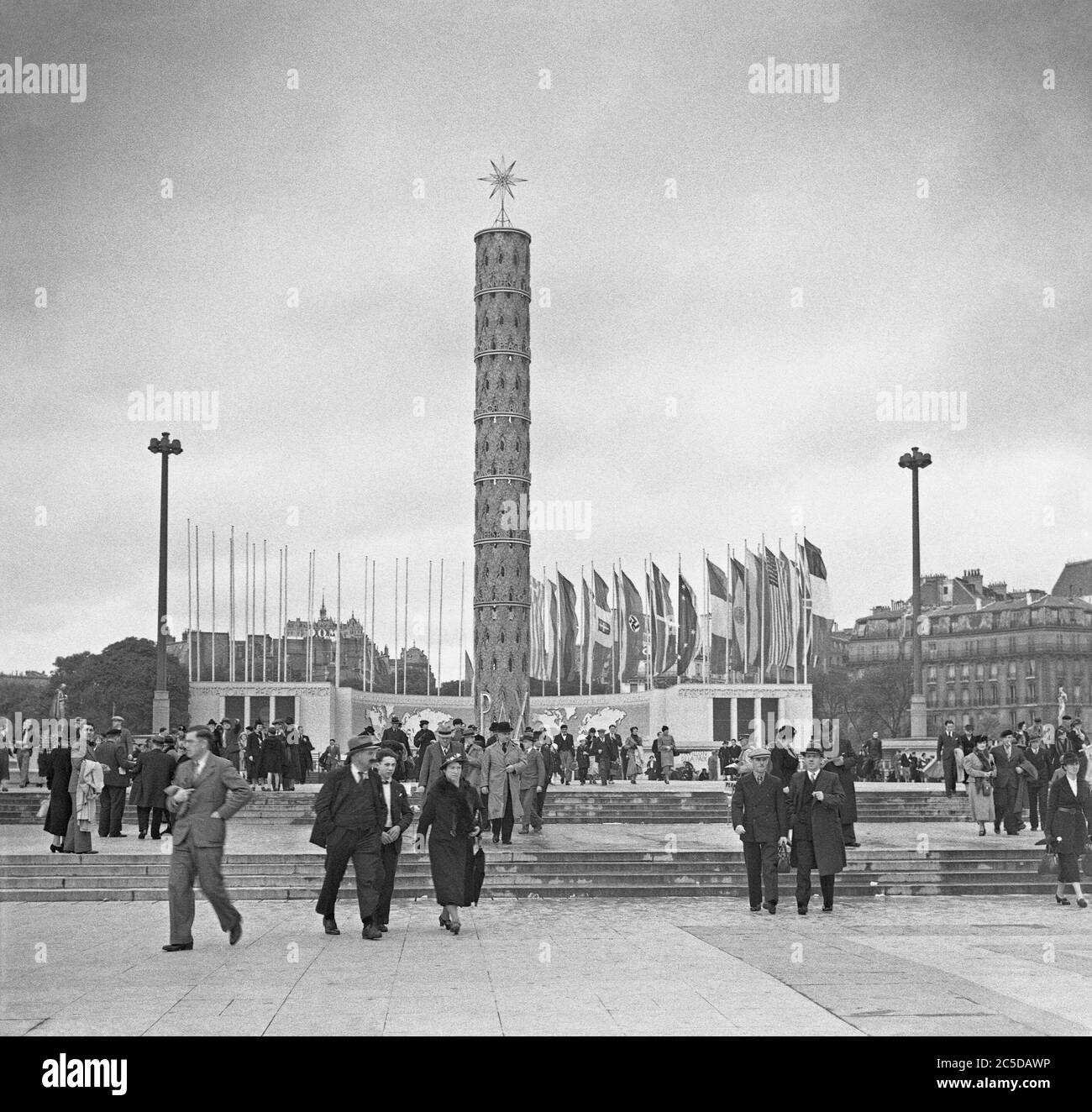 A view of the 1937 Expo (World's Fair or Exposition) held in Paris, France – here people are entering the site at its north end, The Trocadero. The tall tower has the word 'Pax' (Peace) at its base and the flags of nations flutter in the breeze – including the German flag with its swastika. Stock Photo