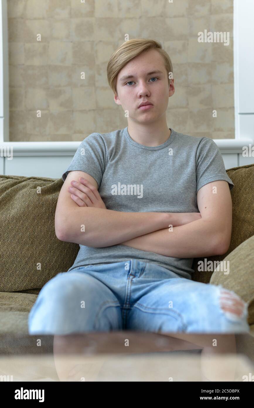 Young handsome blond teenage boy with arms crossed sitting at home ...
