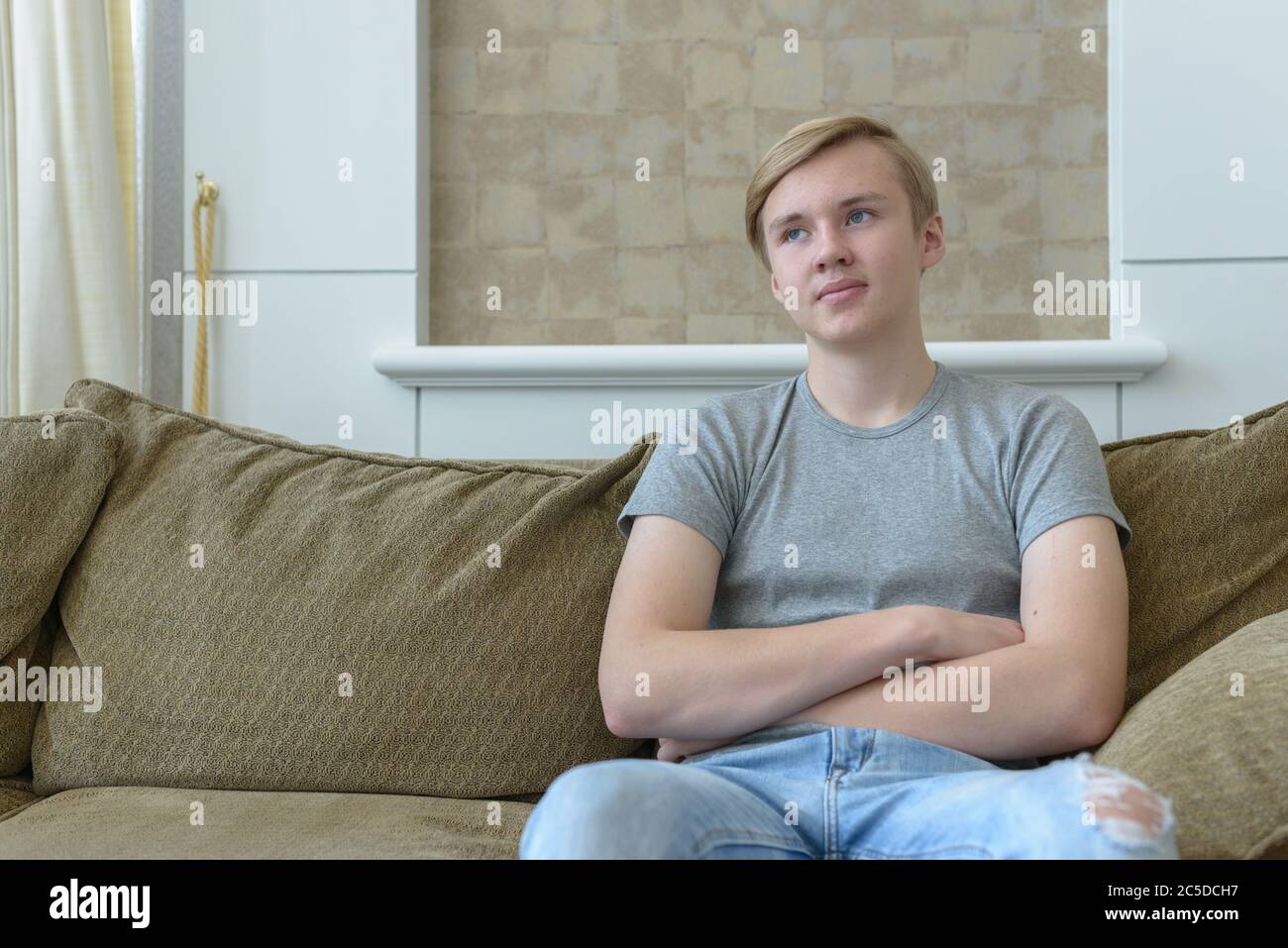 Young handsome blond teenage boy thinking while sitting on the sofa at ...