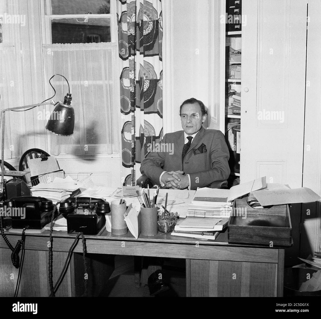 1968, historical, a headmaster of an inner London boys boarding school sitting at his desk, covered completely with paper-work and files, Lambeth, Southeast London, England, UK. An anglepoise lamp and two bakelite telephones of the era are also on the overcrowded desk. Stock Photo