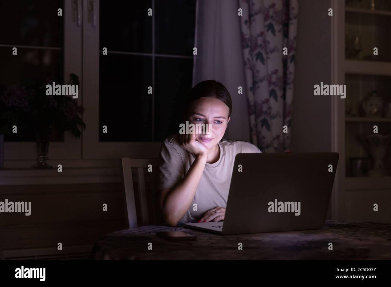 Woman sitting on the table, watching a movie on laptop late at night at home, sharing social media. Stock Photo
