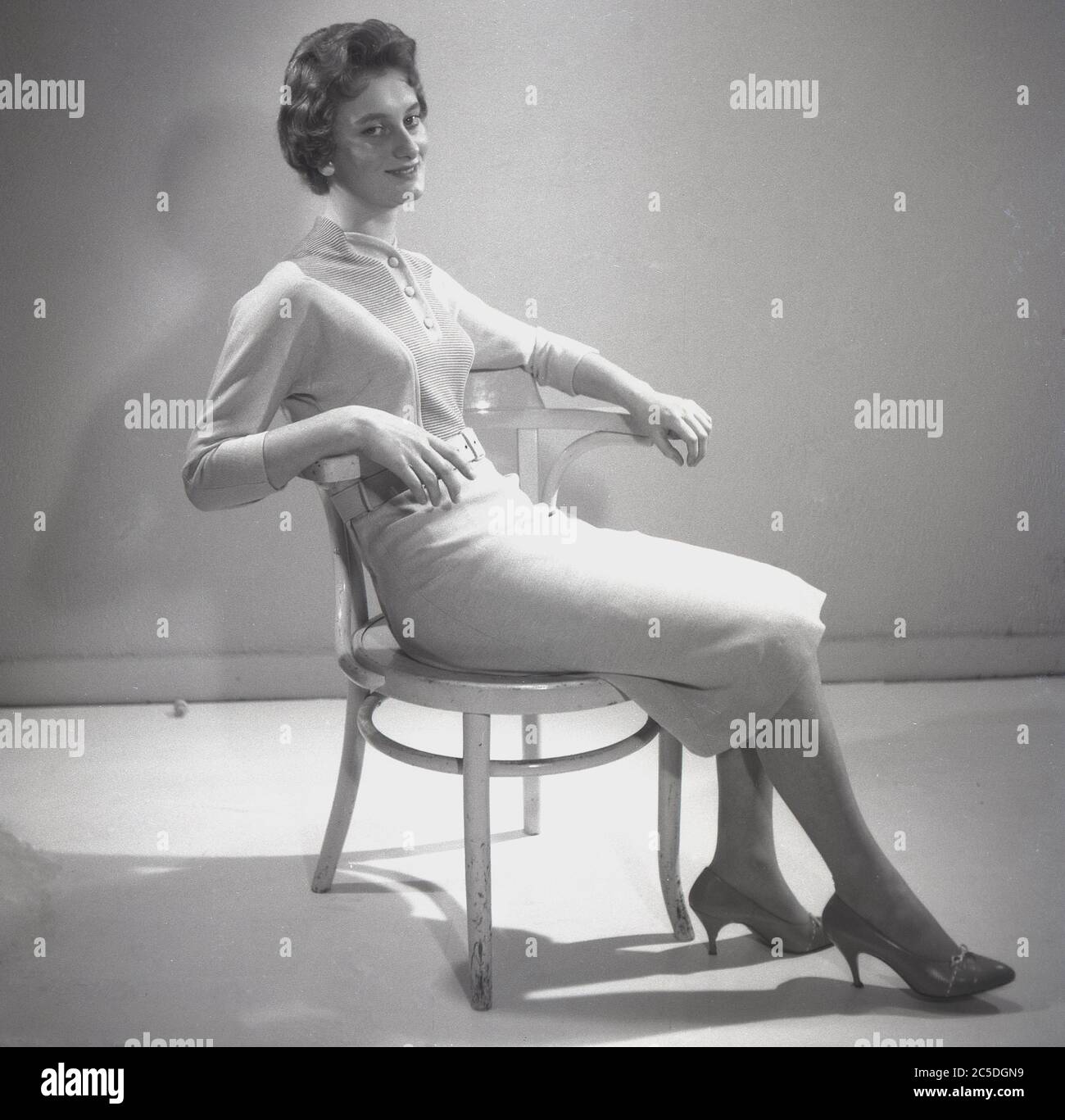 Late 1940s, early '50s, historical, an elegant, graceful English young lady, mid to late twenties in a studio in front of a screen sitting in a wooden button chair modelling the latest female fashions, a two piece outfit of woollen top and skirt, England, UK. In this era, the austerity look of the war years was replaced by the femininity of a softer look, narrow shoulders,, fitted waist, bust emphasis and longer skirts, with influence from Parisian fashion designers such as Christian Dior. Stock Photo