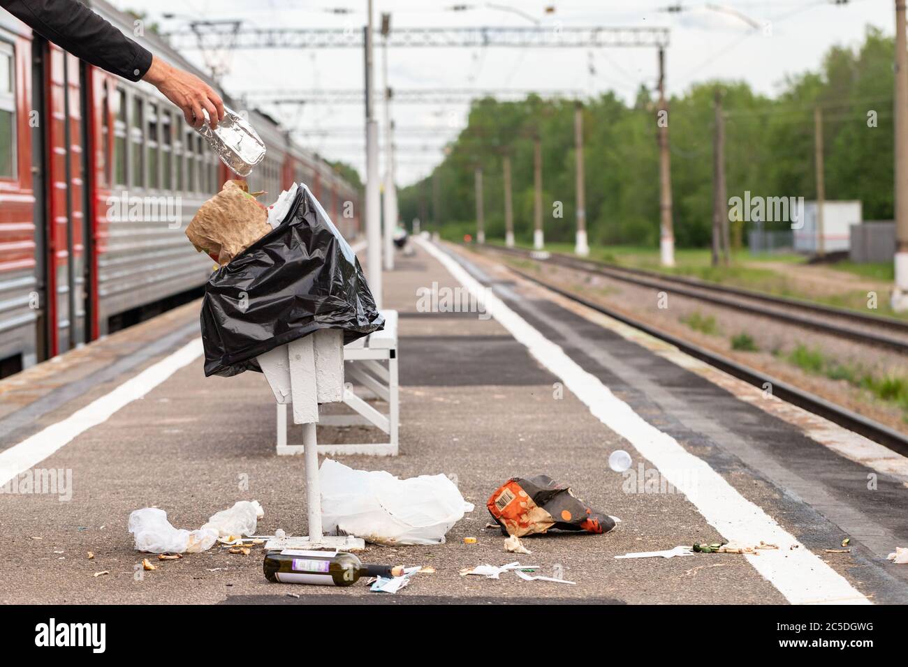 Hand of unknown man throws trash into overcrowded trash basket on a railway platform. Garbage bin. A pile of plastic waste trash on the floor. Stock Photo