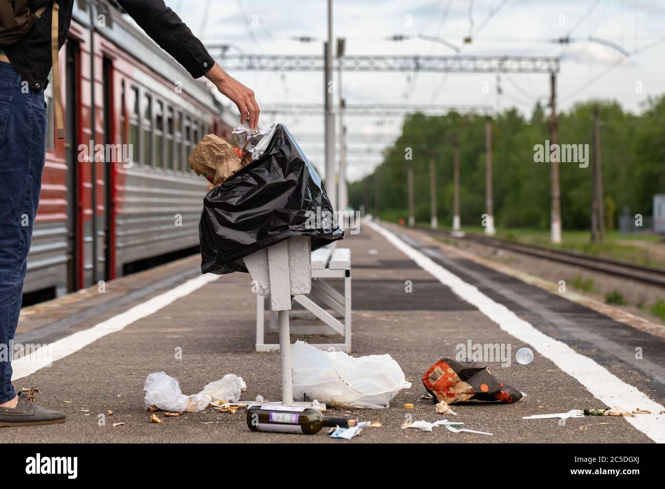 Hand of unknown man throws trash into overcrowded trash basket on a railway platform. Garbage bin. A pile of plastic waste trash on the floor. Stock Photo