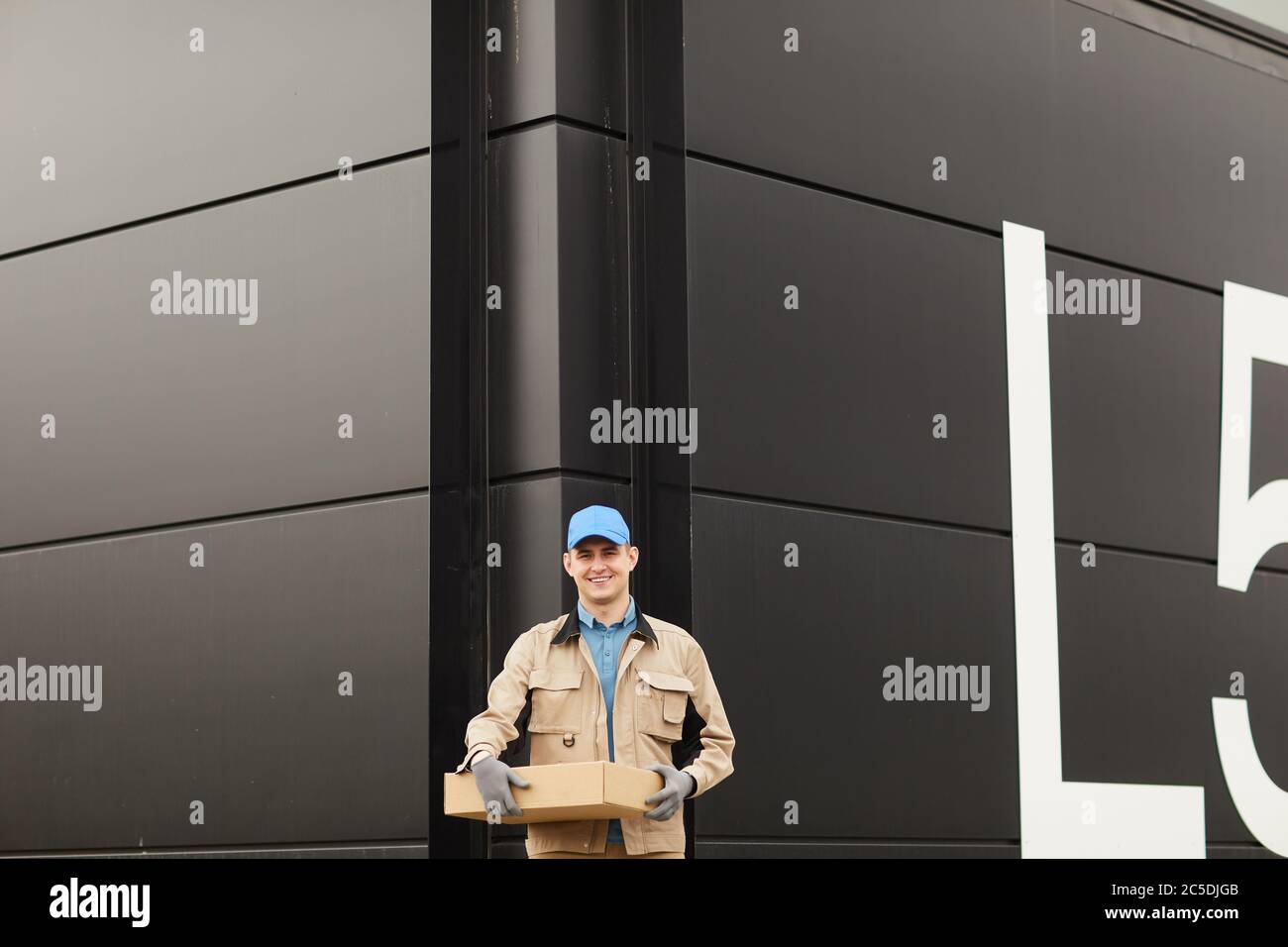 Portrait of young courier in uniform holding parcel and smiling at camera while standing against the warehouse outdoors Stock Photo