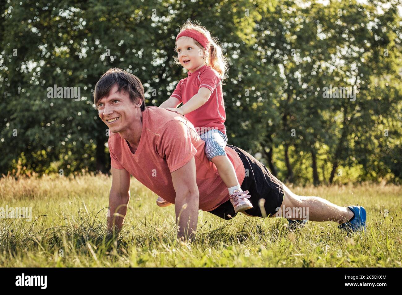 Father playing with his cute young daughter outdoors as he does press-ups on the grass with her riding on his back laughing in an active healthy lifes Stock Photo