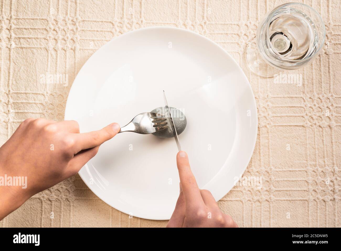 Minimal Easter composition. Hands cutting gray easter egg on white plate with fork and knife on linen tablecloth, glass of water Stock Photo